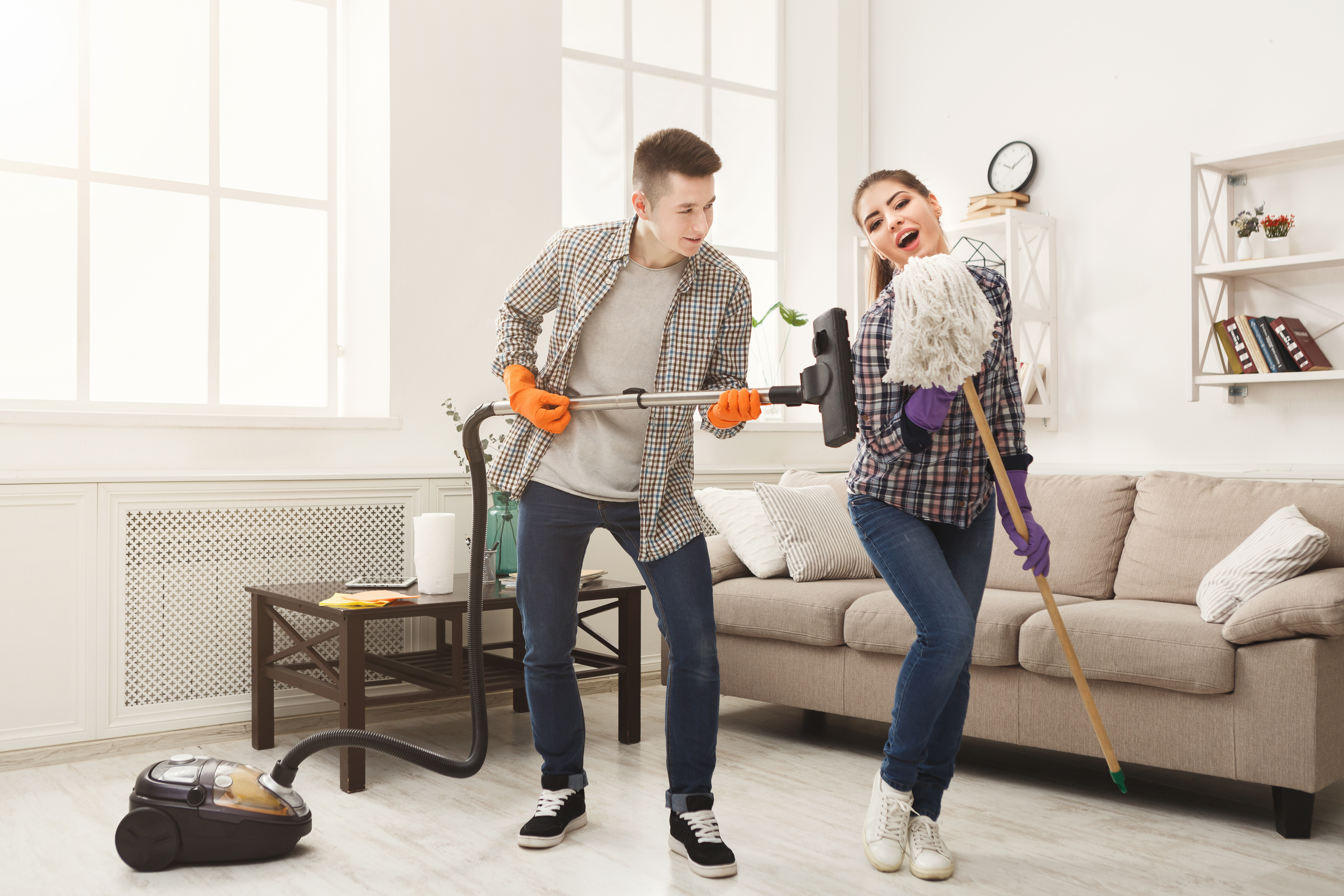 Young couple cleaning home, playing with mop and vacuum cleaner, having fun in living-room. Housekeeping and home cleaning concept